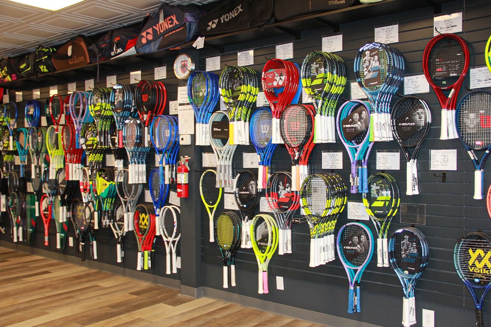 Wall of tennis rackets at Forever Tennis featuring Babolat, Wilson, Yonex, Head, and more.